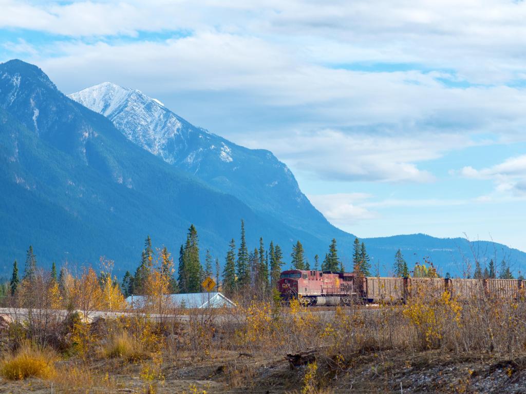 A red freight train passes in front of the rugged Canadian Rocky Mountains near Golden, British Columbia on a cloudy fall day