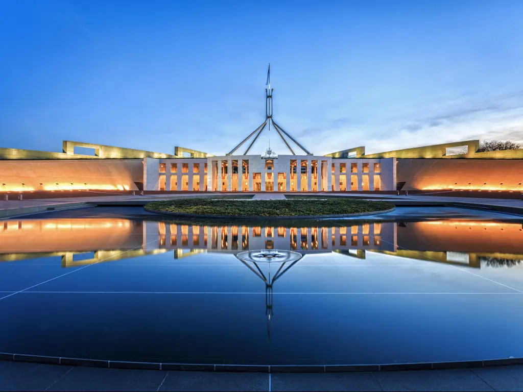 Wide modern white building with tall, narrow windows in central section, with deep blue sky behind. Buildings and sunset reflected into still water in front