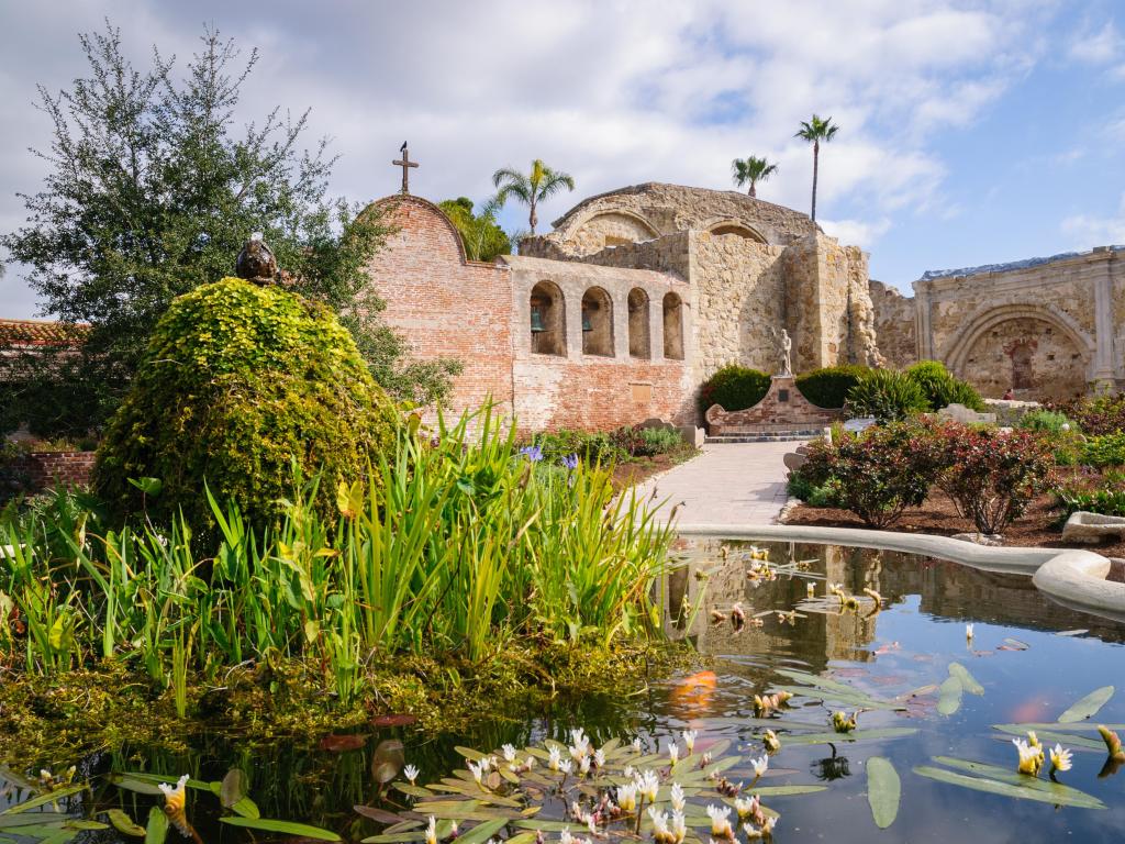 Courtyard of Mission San Juan Capistrano in California on a sunny day with a pond in the foreground and building in the background.