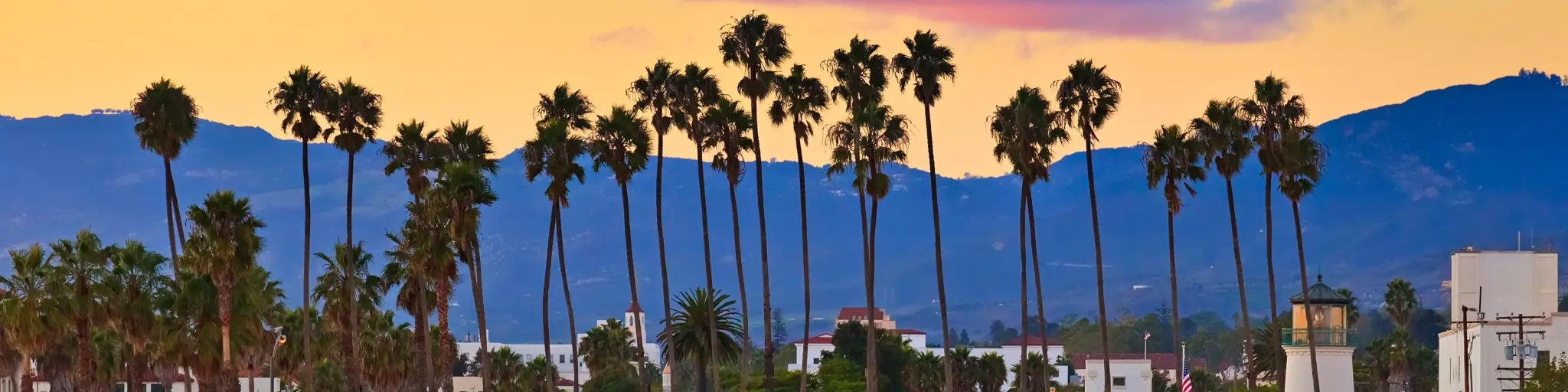 View of the city from the pier, shielded by palm tress with mountains in the background during sunset