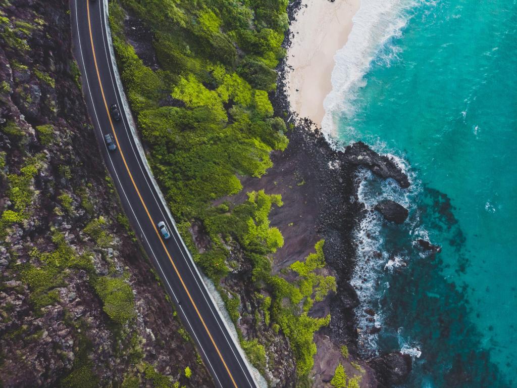 Aerial view of a road running along the coast in Hawaii, with small cars in the distance below